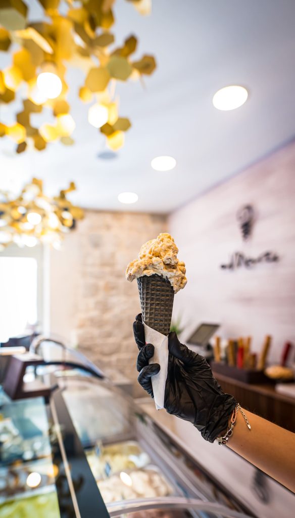 Gloved hand holding black gelato cone with scoop of gelato inside gelateria with aMare logo in the background